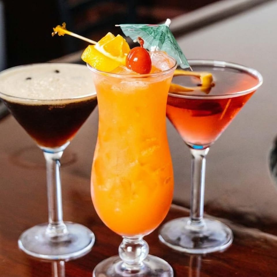 Three colorful cocktails on a polished bar, including an orange fruit-garnished drink, dark coffee cocktail, and red martini.