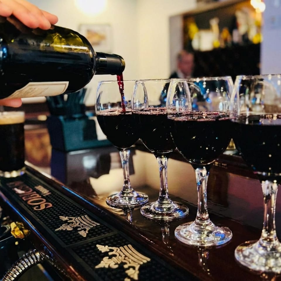 Bartender pours red wine from a bottle into a row of glasses at a warmly lit bar.