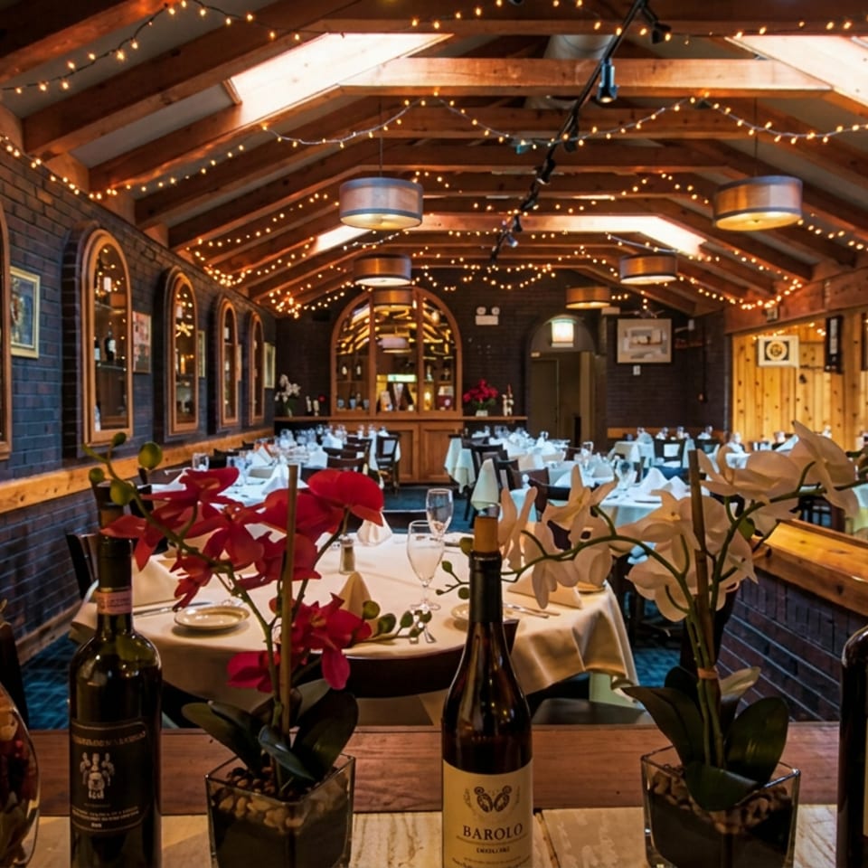 Elegant Italian restaurant dining room with white tablecloths, wine bottles, orchids, exposed beams, and warm string lights.